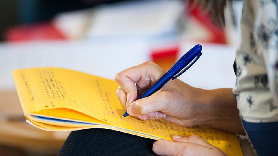 A hand holding a blue pen and writing on a yellow notepad