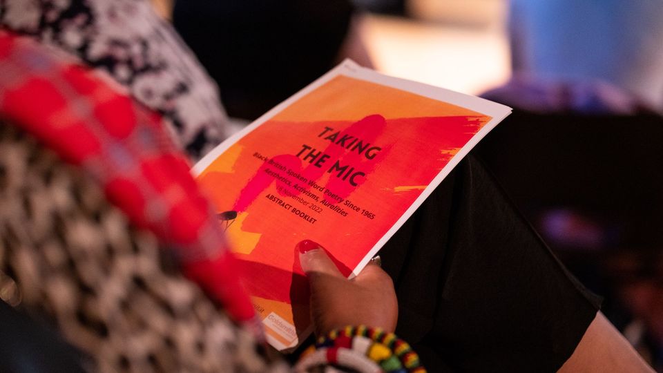 A red paper event programme with the words 'Taking the Mic' is held by an attendee. 