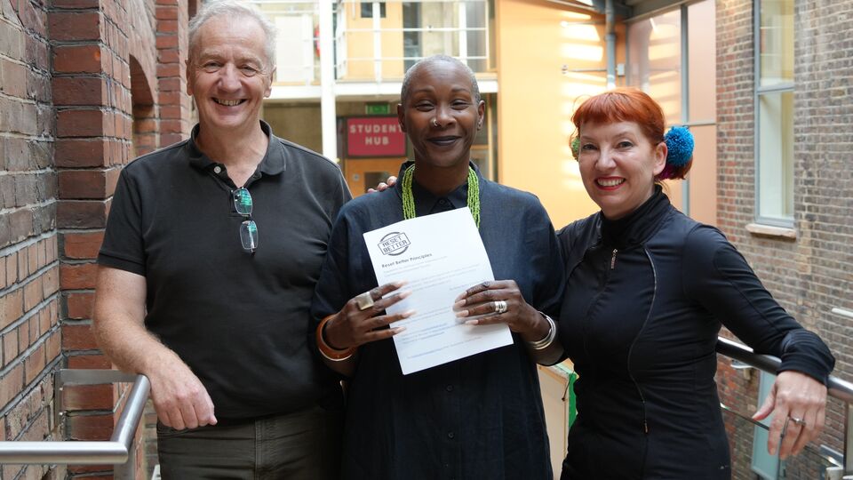 Central's David Ripley and Dr Kathy Sandys stand to either side of Principal Josette Bushell-Mingo OBE who holds a signed copy of the reset better charter. All three wear black and stand in the School's brick walled atrium which is filled with sunlight.