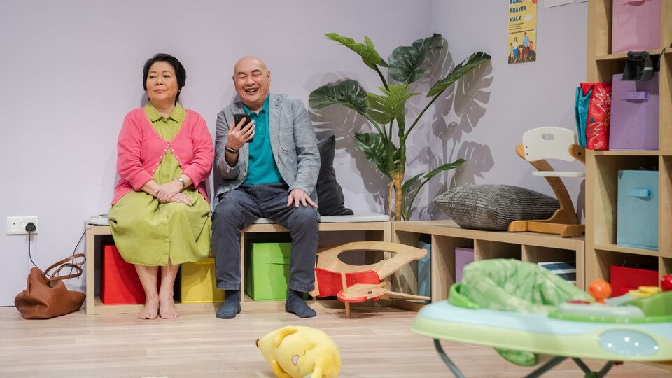 Two actors sit on a bench in a room with assorted toys and brightly coloured boxes in wooden shelves. One is holding a phone and laughing