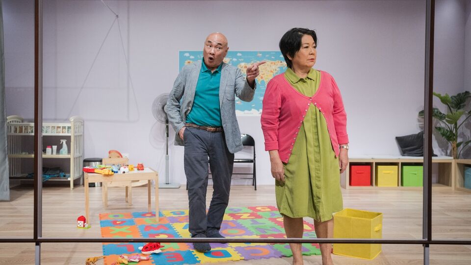 Two actors stand in a room with toys and a mat with the alphabet in coloured letters. One is pointing to the other, who is looking into the distance