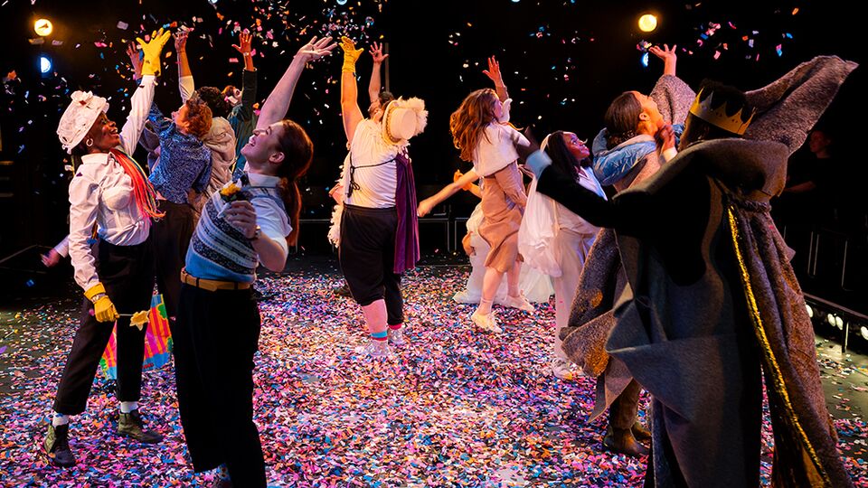 Students on stage in a production raise their arms and throw confetti in celebration