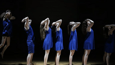 Seven people wearing blue clothes stand in profile to the camera with their arms raised to their heads which look upwards. The farthest person to the left is jumping.