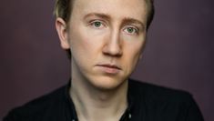 Moody portrait of a blond actor in a dark shirt in front of a dark purple wall.