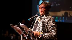 Presenter and poet Carol Leeming MBE FRSA wears red glasses and a leopard print jumper, stands at a music stand in front of a microphone addressing attendees