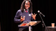 Keynote speaker Kayo Chingonyi FRSL wears a dark blue jumper and white collared shirt, stands at a music stand in front of a microphone addressing attendees