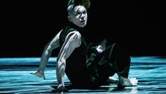 A dark stage hit by dense lines of cold bright light projected from above. A dancer in loose dark trousers and a dark turtleneck vest sits on the floor with bent legs and arms open behind her back.