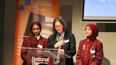 Two Mulberry Arts students in school uniform stand either side of a person giving a talk at a lectern branded with the National Theatre logo