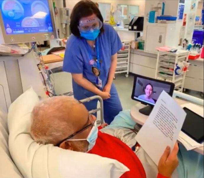 A patient in a hospital bed holding a script, talking to students on a tablet screen with a person in medical scrubs stood next to him to offer support