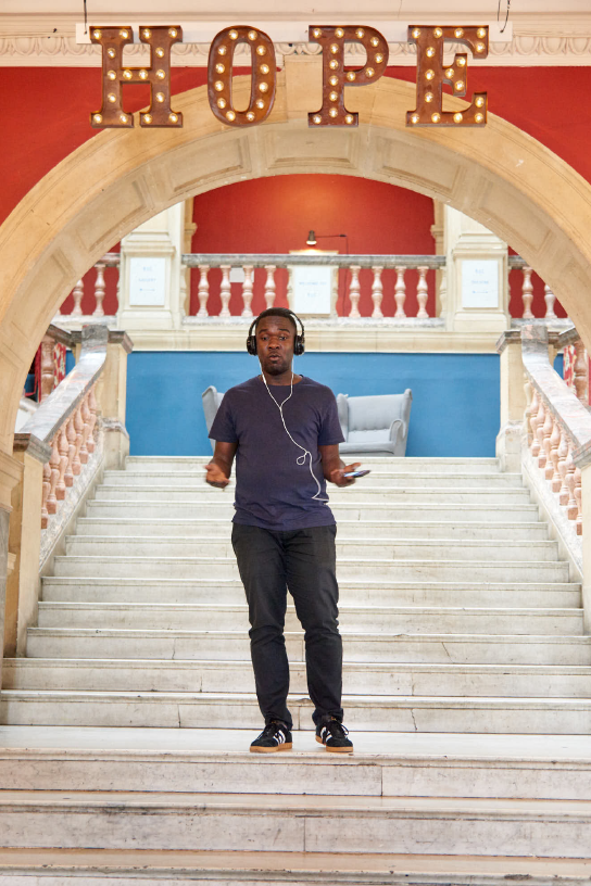 A young black man stands on a set of mob stairs holding the mobile phone and wearing headphones under a sign in titled “HOPE”.