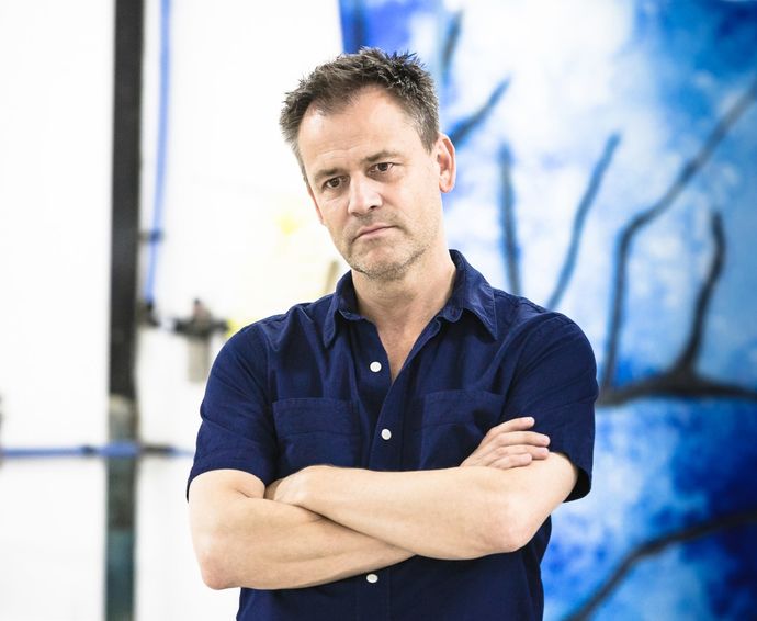 Photo of Michael Grandage CBE wearing a blue, shortsleeved collared shirt, standing against a blue and white wall with his arms crossed in front of his chest