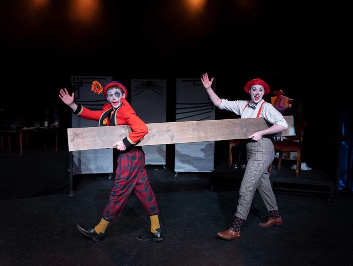 Ryan Denton and Molly Smith dressed in clown costumes and makeup, carry a plank across the stage with one hand raised