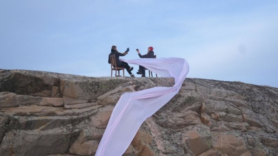 Two people sitting on side of cliff raising glasses, with light pink material swirling in the air
