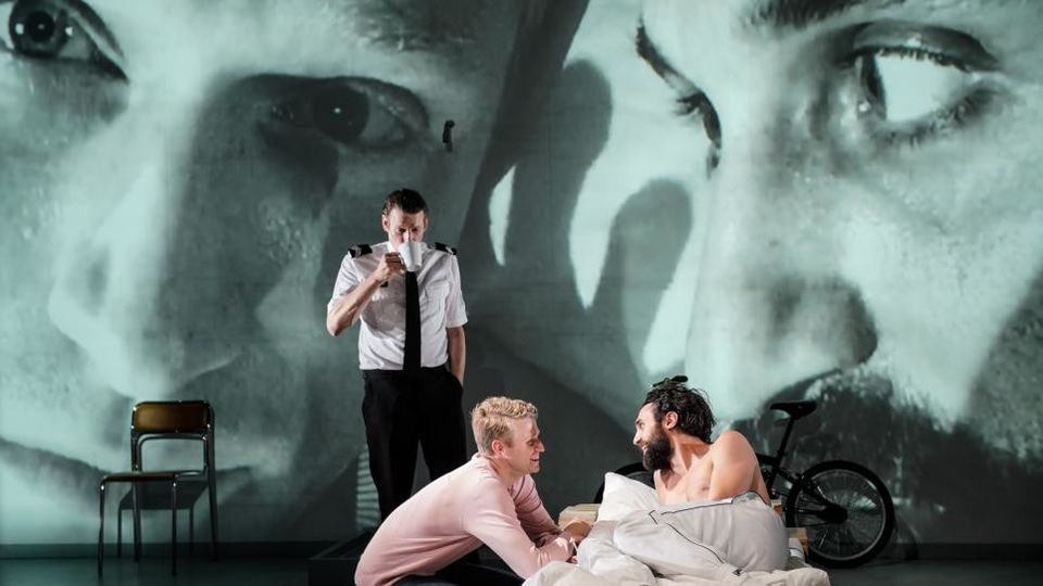 Three actors on stage with two large faces projected on the wall behind them.