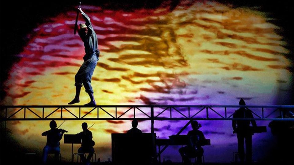 Musicians on stage underneath performer walking across beam with coloured backdrop