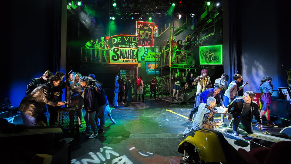 Groups of actors on a stage set that is lit in green and designed to look like a seedy alleyway