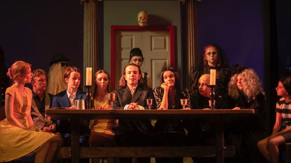Production shot of the cast of Adams Family sat around a table and looking out to the audience