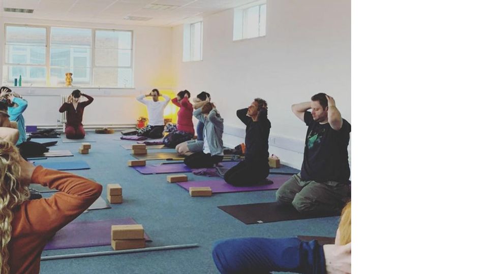 Photo of a movement class showing a group sat in a room on mats with their hands on their heads