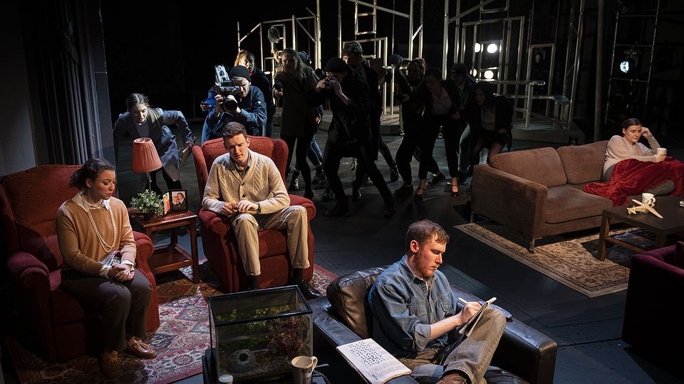 A group of Actors sit on armchairs and sofas in a living room with a group of people in the shadows behind them