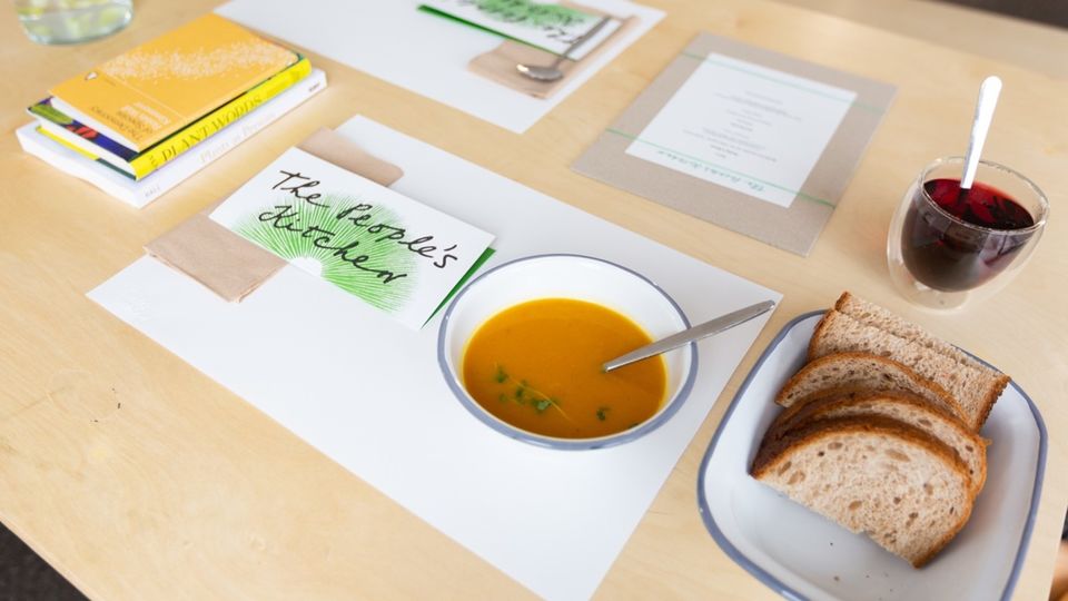 Shot of a table. There is a placemat with a label The People's Kitchen and a bowl of orange soup with a spoon in it on the placemat. Next to the place mat there is an enamel container with five slices of bread in it. A glass of tea is above that and a pile of books on the other side of the placemat. 