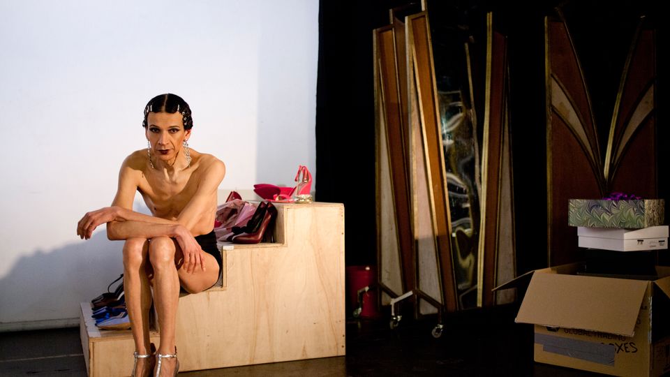 Room with white walls. on the left is a set of plywood steps which have shoes on them and on the right a folded full length mirror and a pile of cardboard boxes. Artist Nando Messias sits on the steps. They have short dark hair, held back either side of a middle parting with diamante clips, long dimante earrings. They are wearing purple pants and silver high heeled t-bar shoes. 
