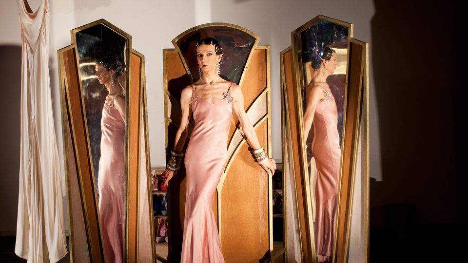 A room with white walls and a mirror with three sections in the middle. Artist Nando Messias stands in front of the middle mirror wearing a pink satin dress. They are reflected in the other two. 