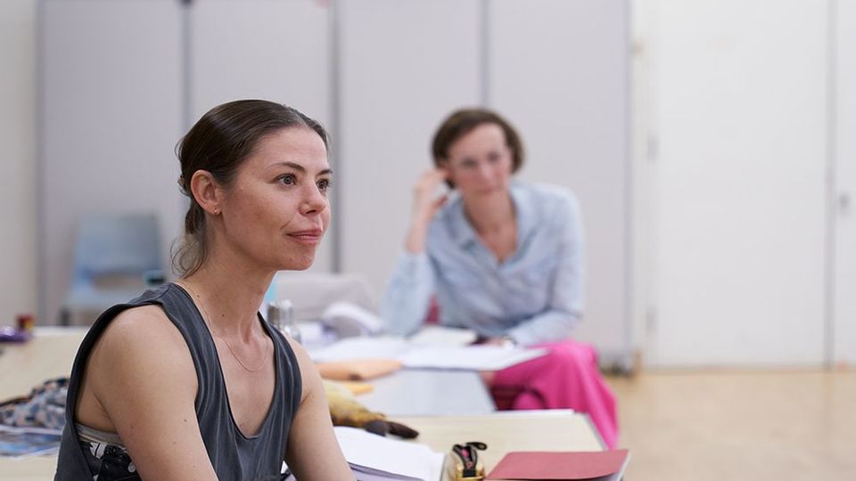 Rebecca Wield is sitting in a rehearsal room at a table, watching activity happening out of frame. Another person is sitting at the table in the background.