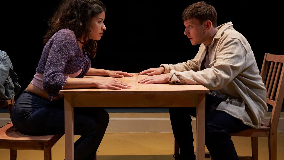 Two actors sit across each other at a table, their hands placed palms down on the tabletop