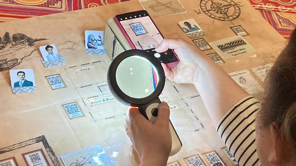A sheet with photographs and other images and questions on it, devising a community map. A person is holding a phone above one of the images and a magnifying glass in the other hand. 