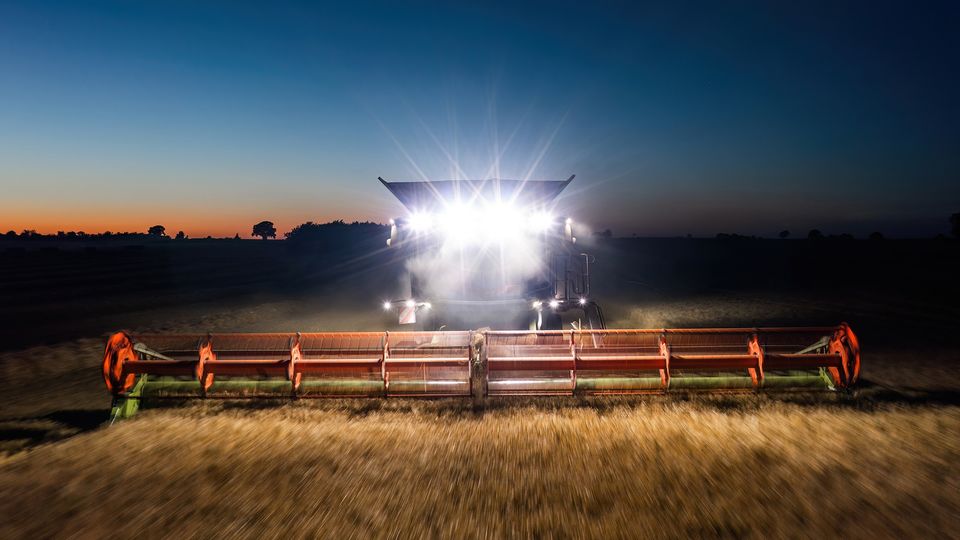 A combine harvester ploughs at night with its lights on. The motion blurs the front of the vehicle