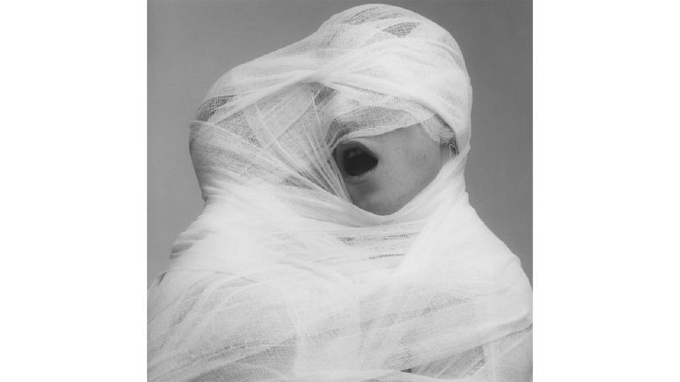 Black and white photography, depicting a couple wrapped in white, blanched gauze, their heads tied together into a cocoon like, sculptural form. One half of the couple has their mouth open and there is no gauze on that part of their face.