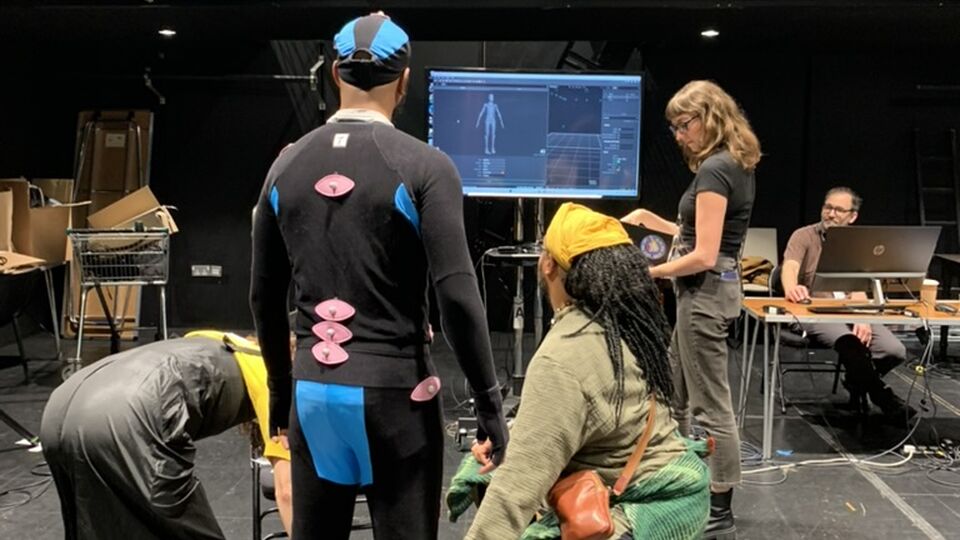Black box studio space. Four people stand in front of a tv screen at eye-level. One of them has motion capture sensors placed on his clothes. 