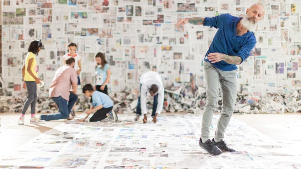  performer performs surrounded by newspaper pages. In the background children interact