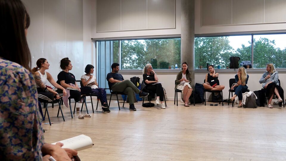 Movement practitioners participating in a discussion, sitting in a large circle inside a rehearsal studio