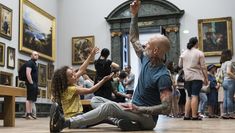 A young girl and a tattooed man dance together in a busy art gallery.