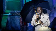 Actor wearing a rain coat and singing while holding an umbrella
