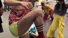 Valerie wearing a bright pink printed top, bright yellow shorts, and red socks dancing in an art gallery with other dancers, audience, and paintings in the background