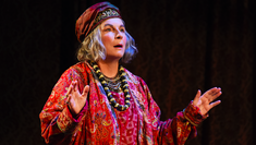 Jennifer Saunders on stage at The Harold Pinter Theatre playing Madame Arcati. She is standing with her mouth ajar with a hat and brightly coloured robe on. 