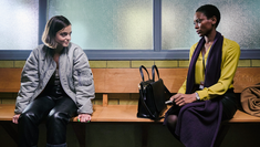Photo of Talitha Campbell (CELINE BUCKENS), Cleo Roberts (TRACY IFEACHOR) in BBC's Showtrail, a whie lady with brown hair in a puffer jacket sits on a bench and smiles at her lawyer, a black lady with short hair in an orange blouse who is also sitting on the bench