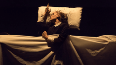 A woman stands in a freestanding bed, her head is tilted to the side on a floating pillow, her arm is resting on a floating sheet. She wears a black suit and her hair is in a bun. 