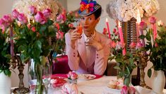 Trans/non-binary individual at table, wearing a hat and pale pink jacket. Pink food and flowers adorning the table