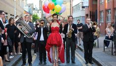 Performer in a red gown, holding a bunch of colourful balloons on the streets with a marching band behind them and audience all around them