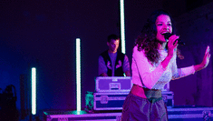 Image of Boadicea Ricketts on a stage singing into a microphone with bright lights in the background and a man standing behind her