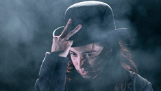 A woman standing on stage with smoke billowing around her clutching at a bowler hat on her head