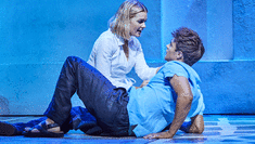 Image of a man and woman together on stage, the woman is leaning over the man looking concerned whilst he's lying down and looking back up at her