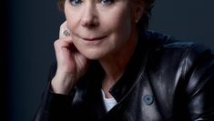 Headshot of Zoe Wanamaker, looking at the camera, wearing a black jacket and sitting against a black background, with her head resting on her right hand