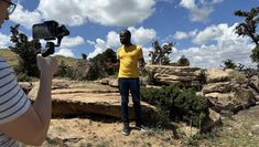 Individual wearing yellow tshirt and blue jeans is being filmed while standing in front of rock formation in Kenya