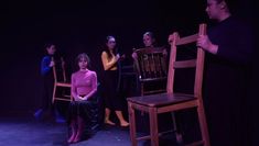 A group of four women move chairs away from one woman sat, singling her out. There is an energy of rejection.