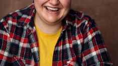 A white woman with short brown hair and green eyes laughing with owl earrings wearing blue jeans, a yellow t-shirt and a blue, white and red plaid shirt in front of a brown background.