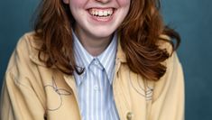 Paige wearing a light button down and yellow jacket smiles into the camera, her red mid-length hair loose. 